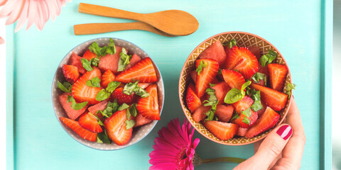 Fruit salad with strawberries, watermelon and basil leaves in ceramic bowls on turquoise wooden tray with female hands. Summer healthy snack
