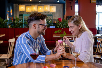 Young romantic couple in a beautiful fancy restaurant. Boyfriend surprise his girlfriend with a red rose and propose to her with an engagement ring.