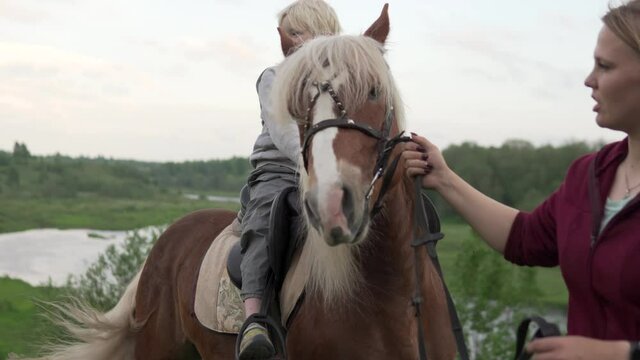 The blonde boy rides a horse in the fields of Novgorod region in the summer.