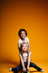 Curly dark-skinned boy rides his little girlfriend on a longboard, and the girl laughs on an orange background