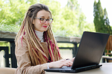 Smiling successful female copywriter with long dreadlocks casual dressed is using black laptop and...