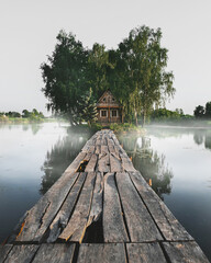 Wooden house in the middle of the lake in the fog. An old wooden bridge leads to a house on the island. Fisherman's house in Stary Solotvyno, Ukraine. Summer morning landscape.