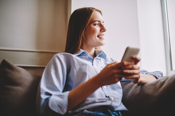 Thoughtful hipster girl in trendy outfit resting after work at comfortable sofa with mobile phone...