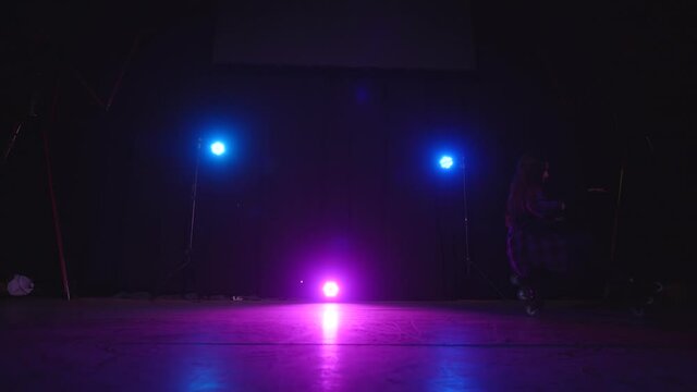Approaching view of pink light of spotlight between blue lamps and a roller-skating girl sitting on front and rear wheels on dark stage. Concept of sport and healthy lifestyle. Casting in the studio.