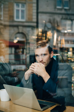 Portrait of businessman with laptop in cafe seen through glass window