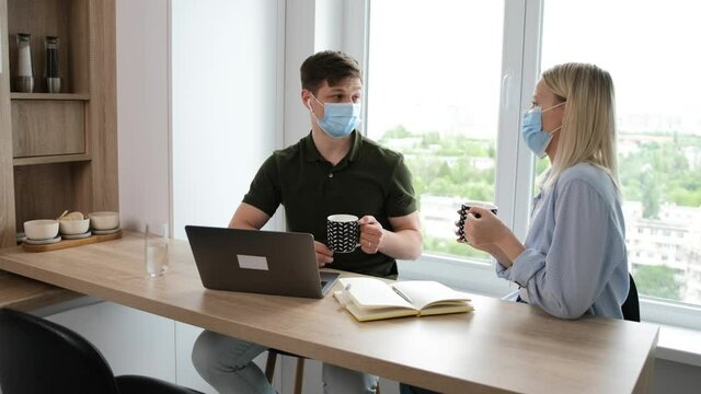 Take a break. Young couple talking about their work day and drink a cup of tea, while they are sitting on kitchen with medicinal mask on face. Stay at home and work from home concept