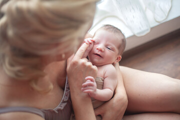 Young mother holding on hands  newborn two weeks baby at home in the room. Home portrait of  happy fаmily against the window