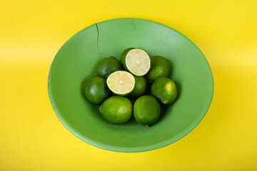 Overview of limes halved and whole in a green wooden bowl on a bright yellow background 
