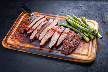 Traditional barbecue sliced dry aged wagyu flank steak offered with green asparagus and chili powder as close-up on a rustic burnt wooden cutting board