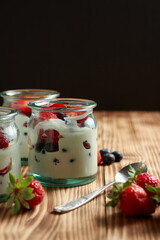 Yogurt pots with berries, strawberries, blueberries and raspberries, on a wooden table with a black background, vertical