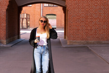 The blonde is walking among the red brick buildings with a coffee in her hands