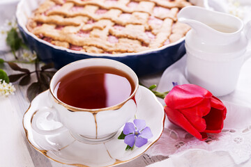 White porcelain cup of tea, and fresh-baked pie. English breakfast still-life with drink and treats red tulip flower and white tablecloth.