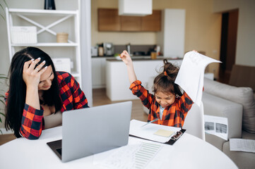Work at home with child. Cute cheerful daughter interferes and distracts her mom while mom try to...