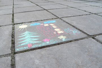 Children's chalk drawing of fir, house, fence, flowers, clouds and sun on square concrete slabs