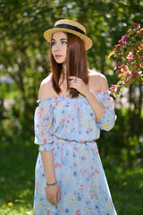 A beautiful girl in a blue dress and a straw hat looks into the distance near a flowering branch of...