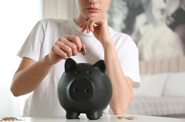 Woman putting money into piggy bank at white table indoors, closeup