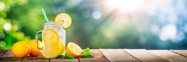 Cold Glass Of Lemonade On Wooden Table With Lemons, Tea Leaves And Sunlight - obrazy, fototapety, plakaty