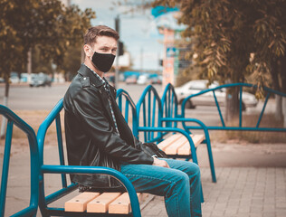 Caucasian sad men sits on the bench and turned his head to the left. He is looking on the photographer.   Dressed into jeans, T-shirt,  jacket and black mask.(Picture has a preset dark colors)