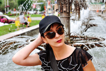 A pretty girl wearing sun glasses and a black baseball cap on background of fountain in summer.
