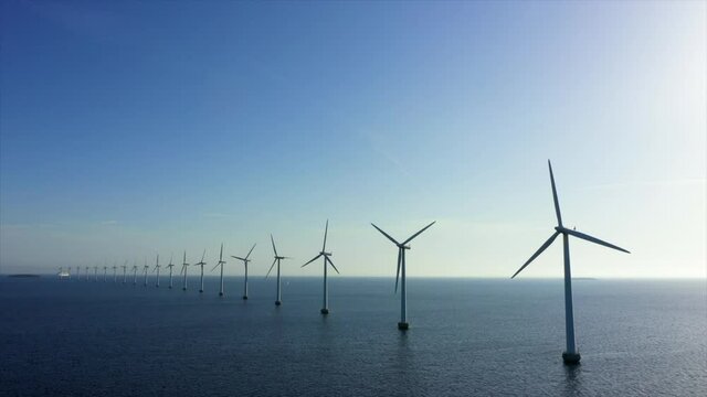 Aerial lockdown shot of wind turbines in sea against blue sky, drone flying over water on sunny day - Copenhagen, Denmark
