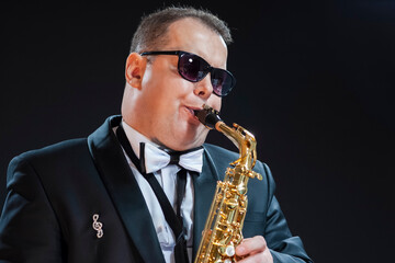 Obraz na płótnie Canvas Music Concepts. Portrait of Passionate and Extravagant Caucasian Saxophonist in Dark Suit Posing in Sunglasses Against Black Background.