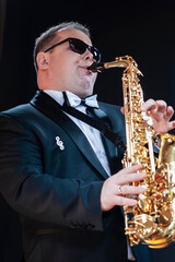 Portrait of Passionate and Extravagant Caucasian Saxophonist in Dark Suit Posing in Sunglasses...