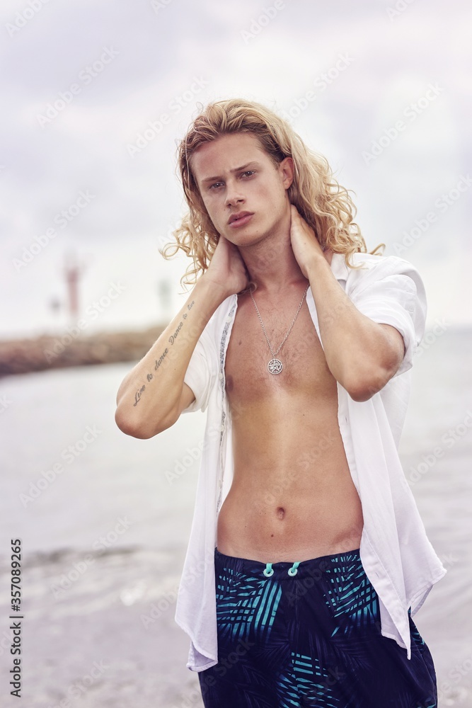 Wall mural vertical shot of a young handsome male with long blond hair wearing an unbuttoned shirt at the beach