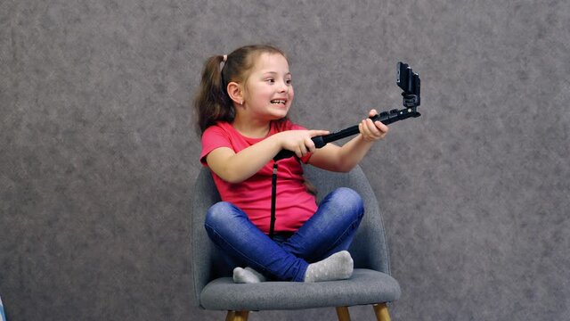 Little girl uses smartphone. Beautiful child in pink t-shirt making selfie with smartphone. Kid sits on the chair and posing on her mobile phone on grey studio background.