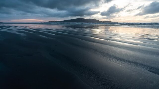 Timelapse at sunset of Kapiti Island on Waikanae beach in New Zealand with reflections of clouds on wet sand