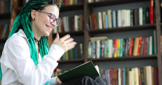 Portrait of laughing girl student with dreads hairs in glasses reads book in library sitting on floor, side view. Girl with green dreadlocks learns literature at University campus. Studying concept.