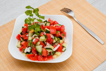 Fresh salad of finely chopped cucumbers, tomatoes and onions with sunflower oil in a white plate and a fork on a wooden table. The concept of healthy nutrition and eco-friendly, vegetarian diet.