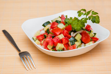 Fresh salad of finely chopped cucumbers, tomatoes and onions with sunflower oil in a white plate and a fork on a wooden table. The concept of healthy nutrition and eco-friendly, vegetarian diet.