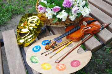 Attributes of the arts. Art palette, brushes, violin, bow, theater mask, a bouquet of roses and jasmine on a garden bench.
