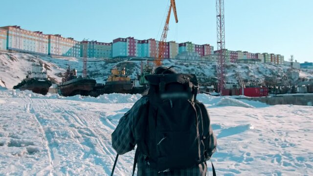 person with backpack walks along frozen sea in local harbour with cranes against city buildings on hilltop in evening backside