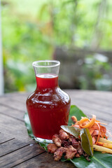 fresh red ginger drink in a glass bottle with shaved secang wood, red ginger, lemongrass, cloves, orange leaves on a wooden table