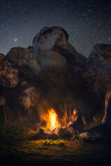 A tourist girl sits by the fire at night among the rocks under a beautiful starry sky. Solitude in the wilderness. Alone with myself.