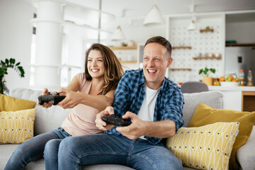 Loving couple playing video games with joysticks in living room. Husband and wife are playing video games at home.  © JustLife