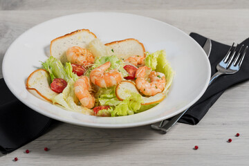 White plate of Caesar salad with shrimps (Ingredients: iceberg Lettuce, crispy croutons, cherry tomatoes, Parmesan cheese and shrimps fried in olive oil) on a light gray background with dark napkins.