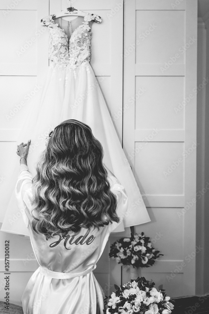 Poster Grayscale shot of a bride wearing a robe and looking at her wedding dress