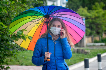 A Caucasian girl in a protective mask stands on an empty street at a bus stop under an umbrella in the spring rain and talks on the phone. Safety and social distance during a coronavirus pandemic.
