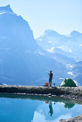 Man enjoying camping in Alps, standing and talking by radio near tent in the rocky alpine mountains near lake with fresh clear water. Concept of beauty nature, hiking and travelling in Alps