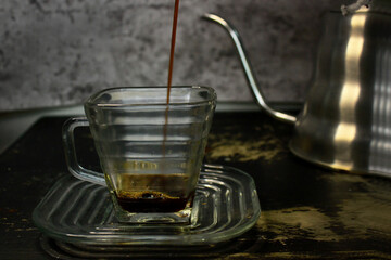 the process of pouring espresso produced mocha pot into a square-shaped coffee cup before mixed with milk, on the table with a swan neck kettle on the back of the glass