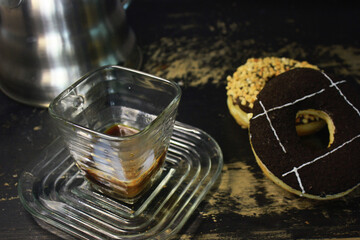 the process of pouring milk on an espresso that is already in a square-shaped coffee cup to make flat white coffee, with a donuts beside it and a swan neck kettle as a background photo