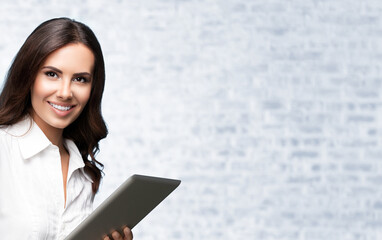 Portrait of happy smiling brunette businesswoman using tablet pc, against white brick wall...