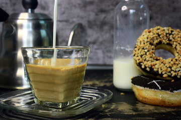 the process of pouring milk on an espresso that is already in a square-shaped coffee cup to make flat white coffee, with a donuts beside it and a swan neck kettle as a background photo