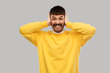 people, noise and stress concept - man in yellow sweatshirt closing ears by hands over grey background