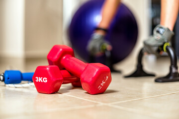 Closeup red dumbbells, at the blurred background blue fitness ball, part of skipping rope and hands...