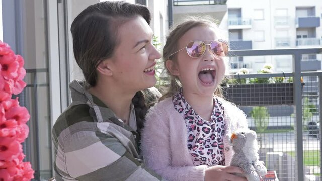 Young woman sits with her six-year-old daughter on the balcony. Mom and chaild are a lot of fun. Photographer is reflected in the mirror glasses of the girl.