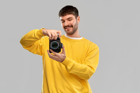 photography and people concept - smiling young male photographer in yellow sweatshirt taking picture with digital camera over grey background