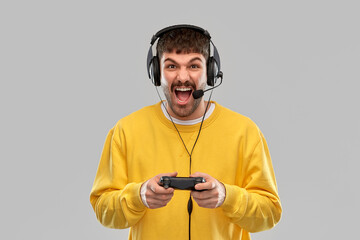 technology, gaming and people concept - angry young man or gamer in headphones with gamepad playing and streaming video game over grey background